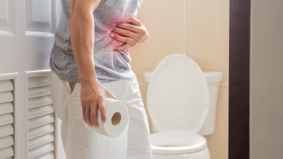 A person holding their stomach in discomfort while standing next to a toilet with a roll of toilet paper